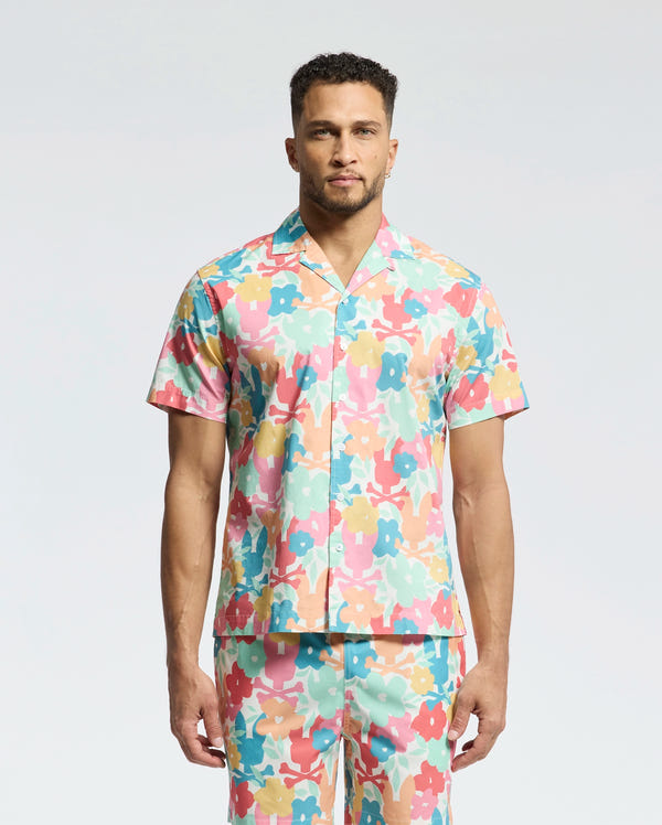 A man faces forward in a Psycho Bunny MENS MEYLON STRETCH POPLIN SHIRT (MT0300034), featuring a colorful floral print, camp collar, and embroidered logo. He wears matching pants; the background is plain light gray.