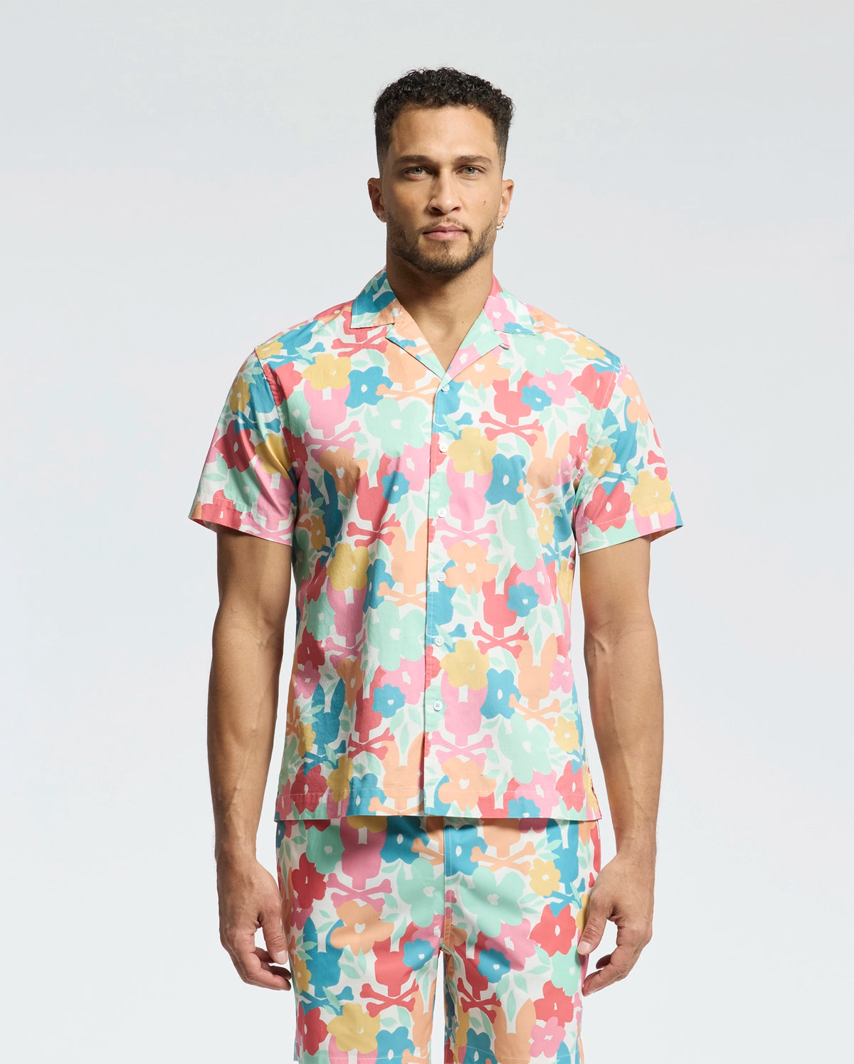 A man faces forward in a Psycho Bunny MENS MEYLON STRETCH POPLIN SHIRT (MT0300034), featuring a colorful floral print, camp collar, and embroidered logo. He wears matching pants; the background is plain light gray.