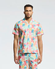 A man faces forward in a Psycho Bunny MENS MEYLON STRETCH POPLIN SHIRT (MT0300034), featuring a colorful floral print, camp collar, and embroidered logo. He wears matching pants; the background is plain light gray.