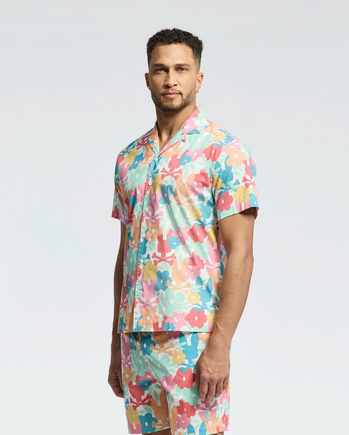 A man stands against a plain background wearing the Psycho Bunny MENS MEYLON STRETCH POPLIN SHIRT - MT0300034, a short-sleeved camp collar shirt with matching shorts, both in a vibrant abstract floral pattern of pink, orange, teal, and white.