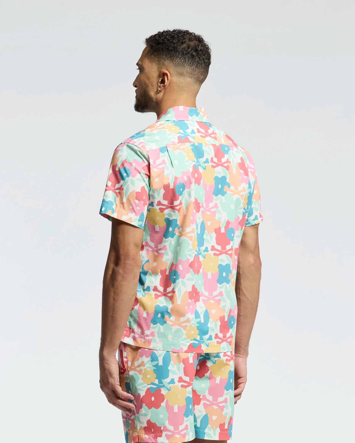 A man stands facing away, wearing the Psycho Bunny MENS MEYLON STRETCH POPLIN SHIRT (MT0300034) and matching shorts, both featuring an abstract floral print in pink, yellow, teal, and white on light gray.