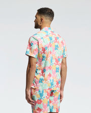 A man stands facing away, wearing the Psycho Bunny MENS MEYLON STRETCH POPLIN SHIRT (MT0300034) and matching shorts, both featuring an abstract floral print in pink, yellow, teal, and white on light gray.