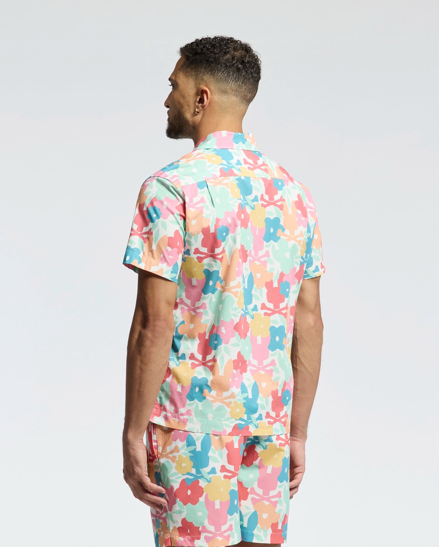 A man stands facing away, wearing the Psycho Bunny MENS MEYLON STRETCH POPLIN SHIRT (MT0300034) and matching shorts, both featuring an abstract floral print in pink, yellow, teal, and white on light gray.