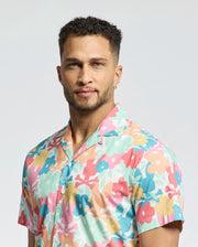 A man with short curly hair, a light beard, and a hoop earring wears the Psycho Bunny MENS MEYLON STRETCH POPLIN SHIRT (MT0300034) with a floral print. He stands against a plain light background, looking calmly at the camera.