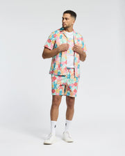 A man wears the Psycho Bunny MENS MEYLON STRETCH POPLIN SHIRT (MT0300034) with a colorful abstract pattern, camp collar, matching shorts, white tee, logo-embroidered socks, and sneakers. He looks aside while holding his shirt collar.
