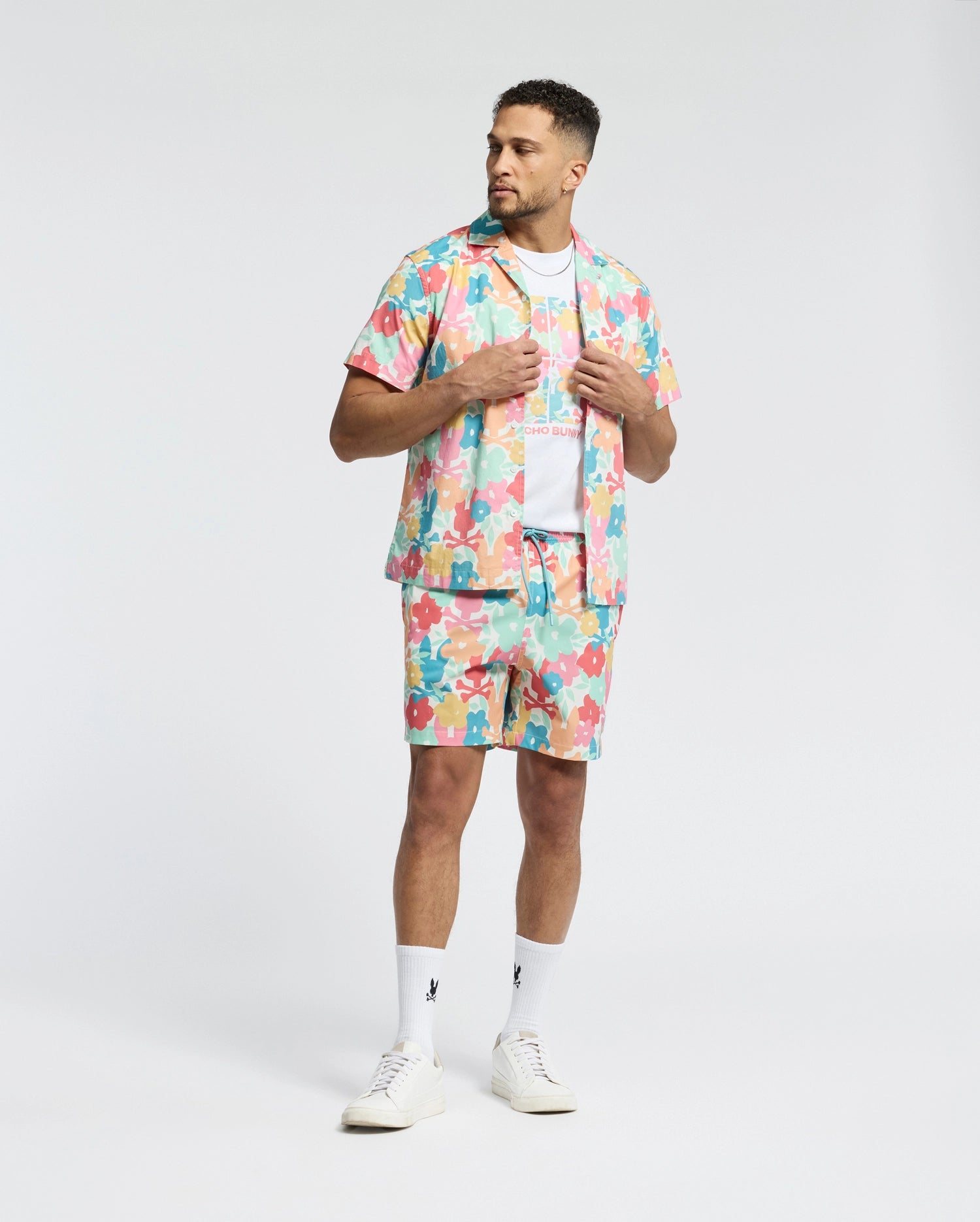 A man wears the Psycho Bunny MENS MEYLON STRETCH POPLIN SHIRT (MT0300034) with a colorful abstract pattern, camp collar, matching shorts, white tee, logo-embroidered socks, and sneakers. He looks aside while holding his shirt collar.