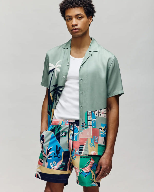 A man stands against a plain background wearing a Psycho Bunny MENS BLAISE TENCEL SHIRT - MT0300056 in mint green with tropical and geometric prints, paired with matching colorful shorts. The relaxed look features 100% Tencel fabric and one hand in his pocket.