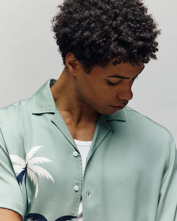 A man with short curly hair wears the Psycho Bunny MENS BLAISE TENCEL SHIRT - MT0300056, made from 100% Tencel with an allover white palm tree print, while looking down against a plain light background.