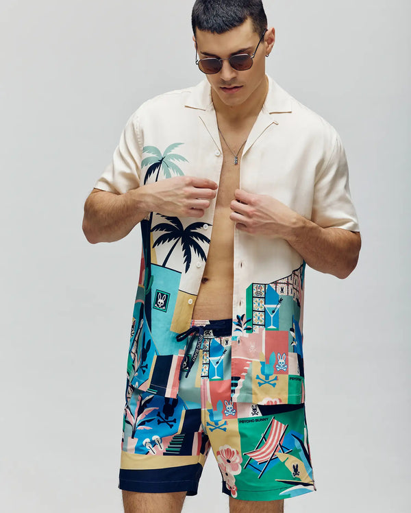 A man wearing sunglasses and a Psycho Bunny MENS BLAISE TENCEL SHIRT - MT0300056 with palm tree tropical designs, paired with matching colorful swim shorts, stands against a plain background.