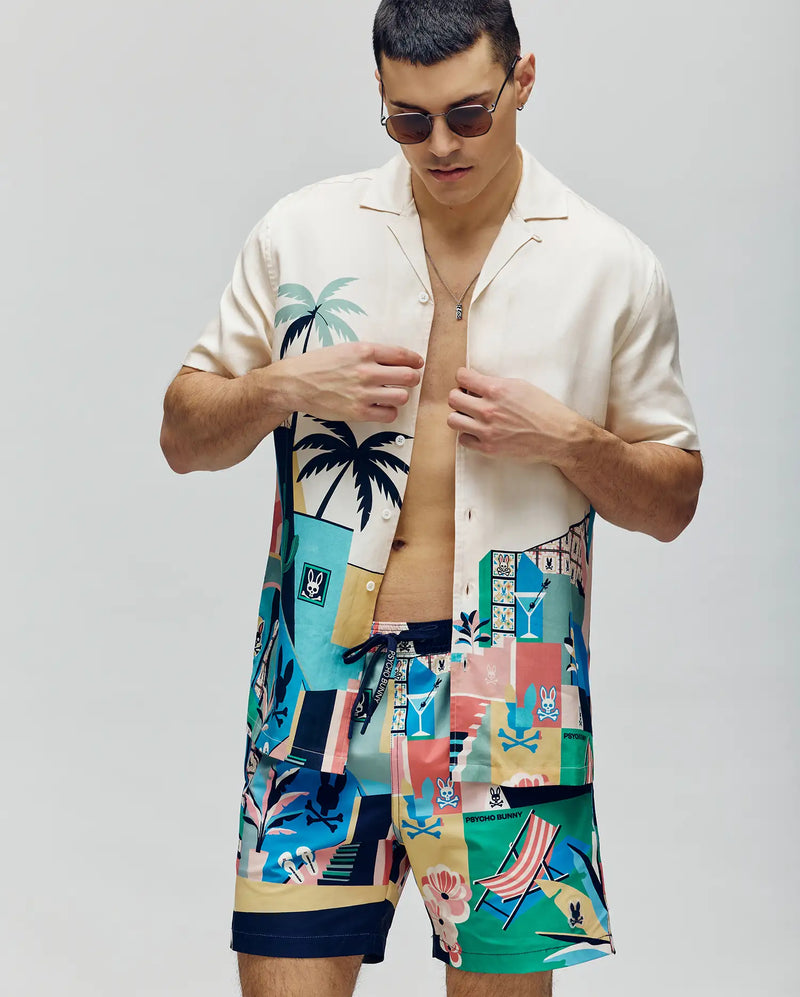 A man wearing sunglasses and a Psycho Bunny MENS BLAISE TENCEL SHIRT - MT0300056 with palm tree tropical designs, paired with matching colorful swim shorts, stands against a plain background.