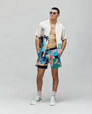 A man in sunglasses, a Psycho Bunny MENS BLAISE TENCEL SHIRT - MT0300056 with a tropical print, colorful shorts, white sneakers, and beige socks stands against a plain light background with his hands in his pockets.