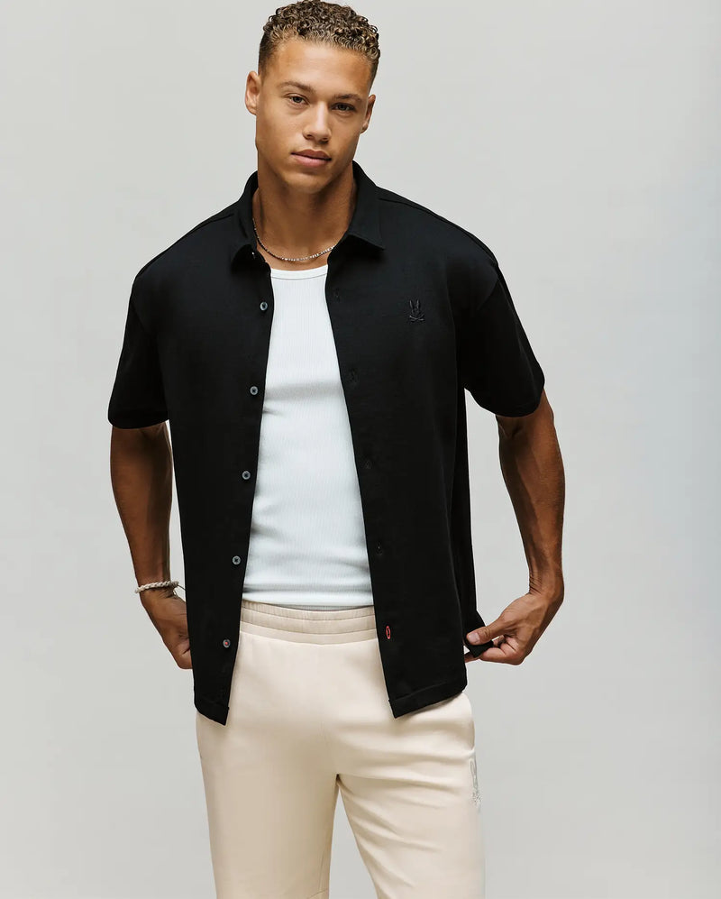 A young man with short, curly hair wears the Psycho Bunny MENS KIAN OVERSIZED FIT SHIRT - MT0300063 in black over a white tank top and cream pants, standing against a plain light background.
