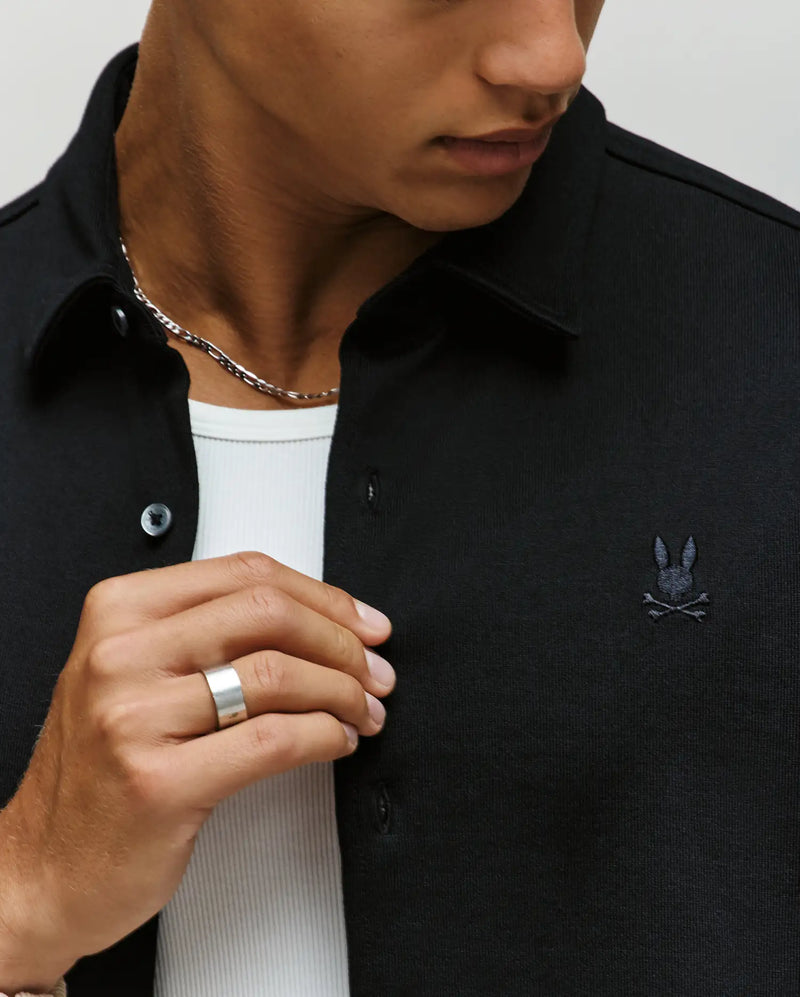 A man wears the Psycho Bunny MENS KIAN OVERSIZED FIT SHIRT (MT0300063) in black, featuring mother-of-pearl buttons and a small bunny logo, styled over a white ribbed shirt with a chunky silver ring and chain necklace, shown shoulders to mid-chest.