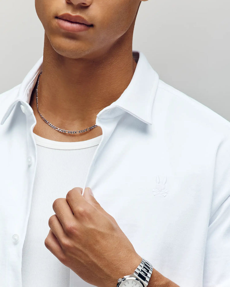 A person in the Psycho Bunny MENS KIAN OVERSIZED FIT SHIRT (MT0300063), a relaxed white button-up with mother-of-pearl buttons, layered over a ribbed tank, accessorized with a silver chain and watch, holding the collar; face partially visible.
