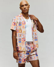 A person with short blond hair showcases the Psycho Bunny MENS SUNDEW ALL OVER PRINT SHORT SLEEVE SHIRT (MT0300064) in vibrant cotton, styled with matching shorts and a white tee, standing against a plain gray background.