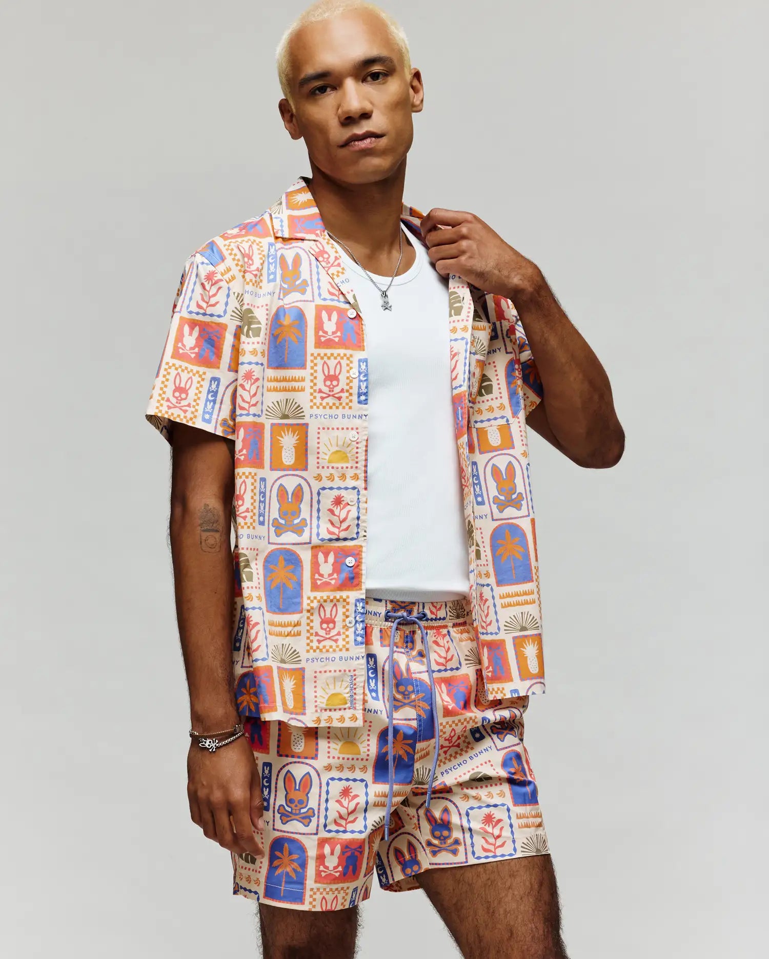 A person with short blond hair showcases the Psycho Bunny MENS SUNDEW ALL OVER PRINT SHORT SLEEVE SHIRT (MT0300064) in vibrant cotton, styled with matching shorts and a white tee, standing against a plain gray background.