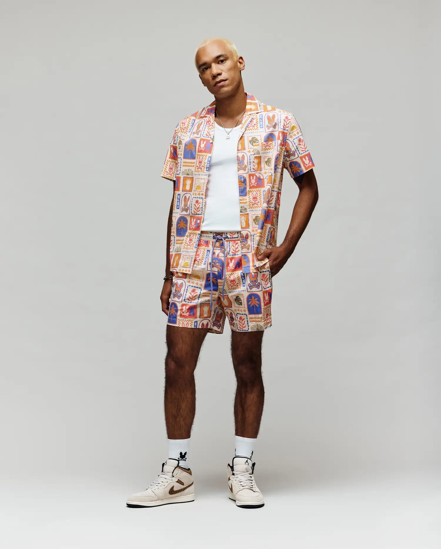 A person with short bleached blond hair wears the Psycho Bunny MENS SUNDEW ALL OVER PRINT SHORT SLEEVE SHIRT (MT0300064), paired with a matching set, white tank top, and white high-top sneakers, standing against a plain light background.