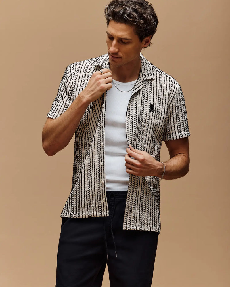 A man wears the Psycho Bunny MENS DEAN CROCHET SHORT SLEEVE SHIRT (MT0300107), a premium cotton blend black-and-white striped shirt with an open collar, layered over a white tee and paired with black pants, adjusting his collar against a beige background.