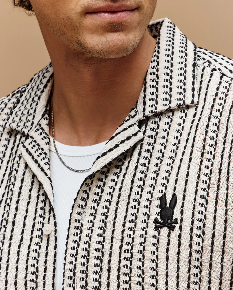 A man wears the Psycho Bunny MENS DEAN CROCHET SHORT SLEEVE SHIRT (MT0300107), a premium cotton blend white shirt with black stripes, open collar, two silver chains, and a black embroidered bunny and crossbones logo. His lower face is visible.