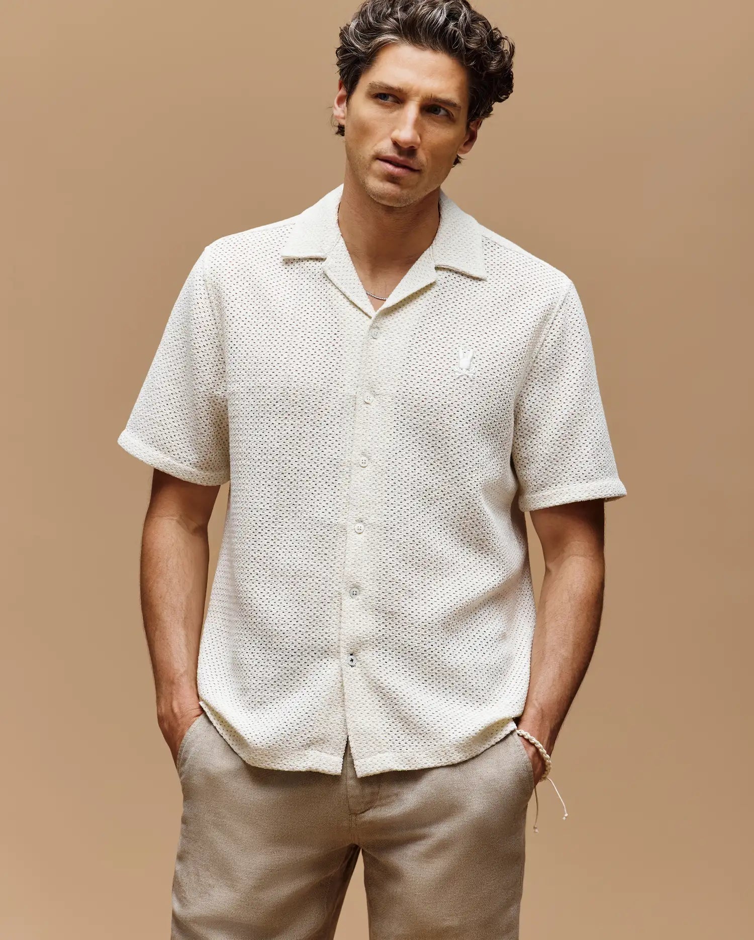 A man stands against a plain beige background, wearing the Psycho Bunny MENS SAMMY CROCHET SHORT SLEEVE SHIRT - MT0300108 in cream and light beige pants, with his hands in his pockets and a neutral expression.