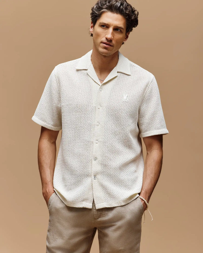 A man stands against a plain beige background, wearing the Psycho Bunny MENS SAMMY CROCHET SHORT SLEEVE SHIRT - MT0300108 in cream and light beige pants, with his hands in his pockets and a neutral expression.