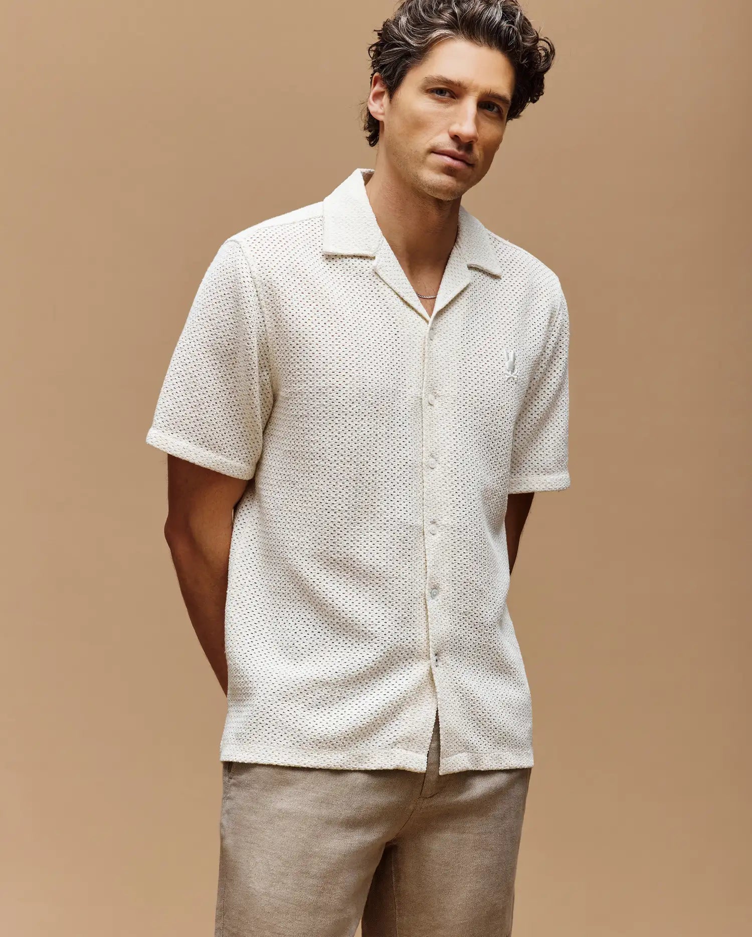 A man with wavy brown hair wears the Psycho Bunny MENS SAMMY CROCHET SHORT SLEEVE SHIRT (MT0300108) in cream and light brown pants, standing with his hands behind his back against a neutral beige background.