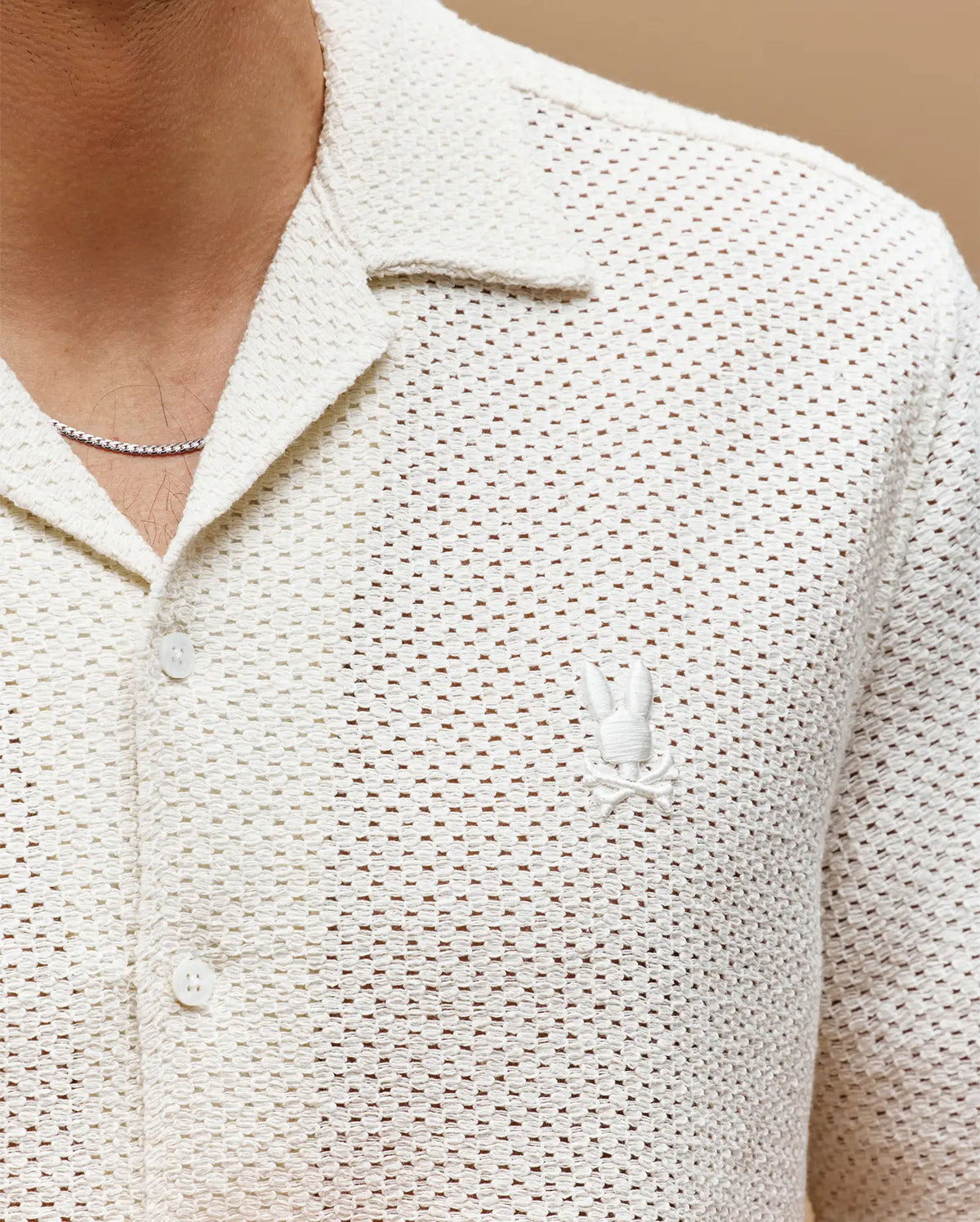 A close-up of a person wearing Psycho Bunny’s MENS SAMMY CROCHET SHORT SLEEVE SHIRT (MT0300108), a cream cotton-blend button-up with a collar and an embroidered Bunny logo on the left chest, paired with a thin chain necklace.