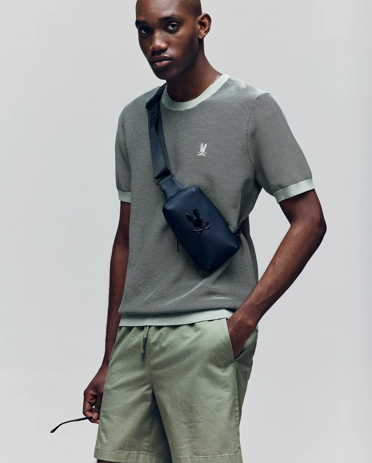 A person wearing the Psycho Bunny MENS MARIO STRIPED SWEATER T-SHIRT (MT0400009) in sage green with matching shorts, accessorized with a black crossbody bag, stands with one hand in their pocket and looks at the camera.