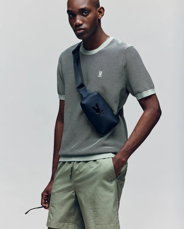 A person wearing the Psycho Bunny MENS MARIO STRIPED SWEATER T-SHIRT (MT0400009) in sage green with matching shorts, accessorized with a black crossbody bag, stands with one hand in their pocket and looks at the camera.
