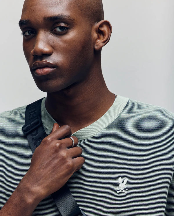 A person with a shaved head wears the Psycho Bunny MENS MARIO STRIPED SWEATER T-SHIRT (MT0400009), featuring light green stripes and an embroidered logo, with a black strap across their chest, gazing slightly to the side.