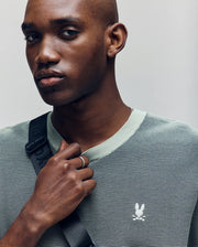 A person with a shaved head wears the Psycho Bunny MENS MARIO STRIPED SWEATER T-SHIRT (MT0400009), featuring light green stripes and an embroidered logo, with a black strap across their chest, gazing slightly to the side.