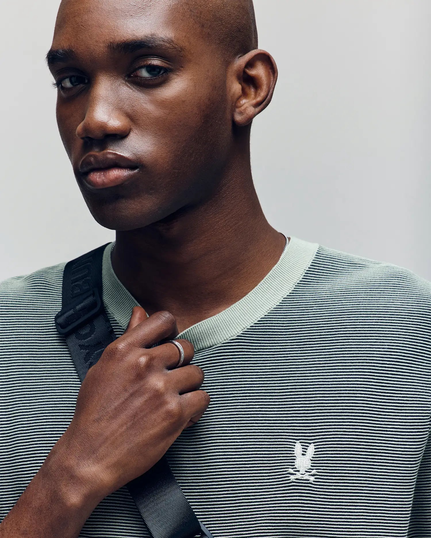 A person with a shaved head wears the Psycho Bunny MENS MARIO STRIPED SWEATER T-SHIRT (MT0400009), featuring light green stripes and an embroidered logo, with a black strap across their chest, gazing slightly to the side.