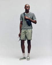 A man wears the Psycho Bunny MENS MARIO STRIPED SWEATER T-SHIRT (MT0400009) with matching shorts, light green sneakers, beige socks, and a black crossbody bag, holding sunglasses against a plain, light background.