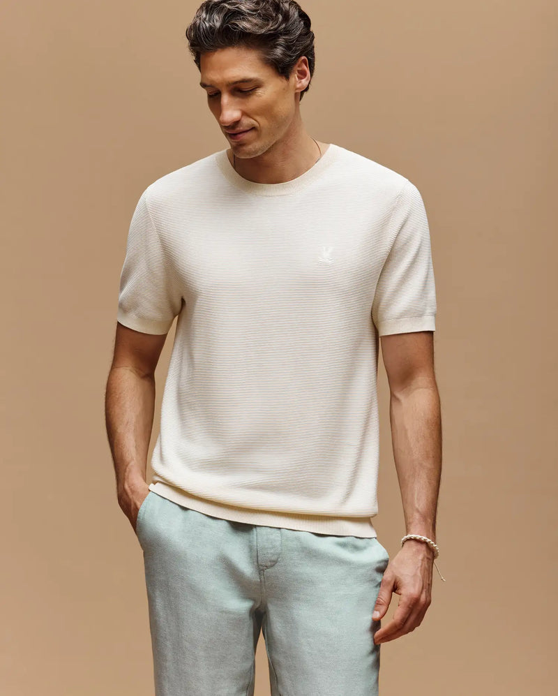 A man wearing the Psycho Bunny MENS MARIO STRIPED SWEATER T-SHIRT (MT0400009), a cream short-sleeve tee with an embroidered logo, and light gray pants stands against a beige background, looking down with one hand in his pocket.