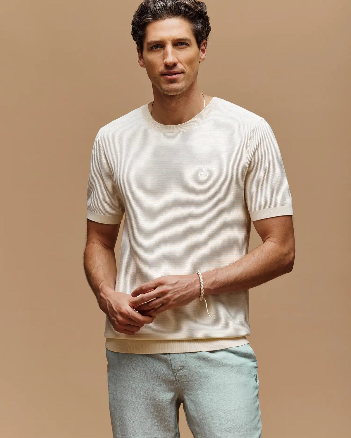 A man with short dark hair wears the Psycho Bunny MENS MARIO STRIPED SWEATER T-SHIRT (MT0400009) and light gray pants, standing against a beige background facing the camera with a relaxed expression.