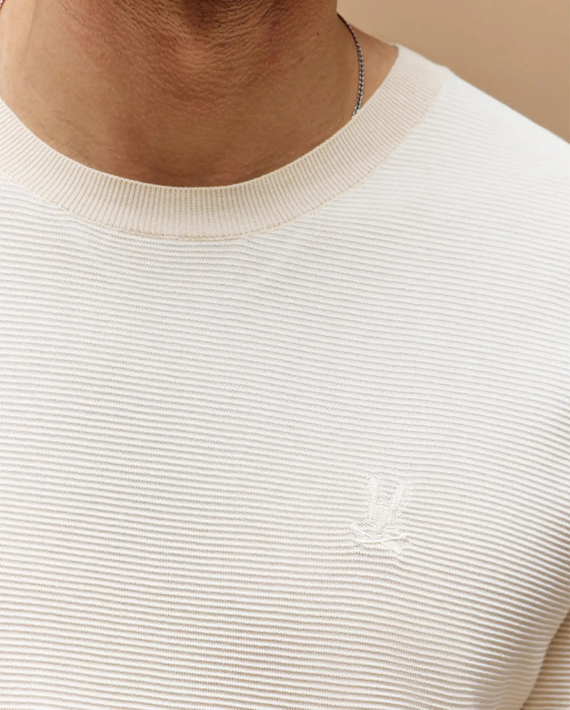Close-up of a person wearing the Psycho Bunny MENS MARIO STRIPED SWEATER T-SHIRT (MT0400009) in light beige with ribbed texture, subtle embroidered logo on the left chest, and a thin chain necklace. Face not visible.