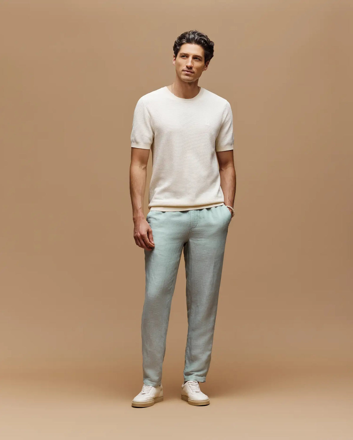 A man stands against a tan background wearing the Psycho Bunny MENS MARIO STRIPED SWEATER T-SHIRT (MT0400009), light blue pants, and white sneakers, with one hand in his pocket and a relaxed expression.