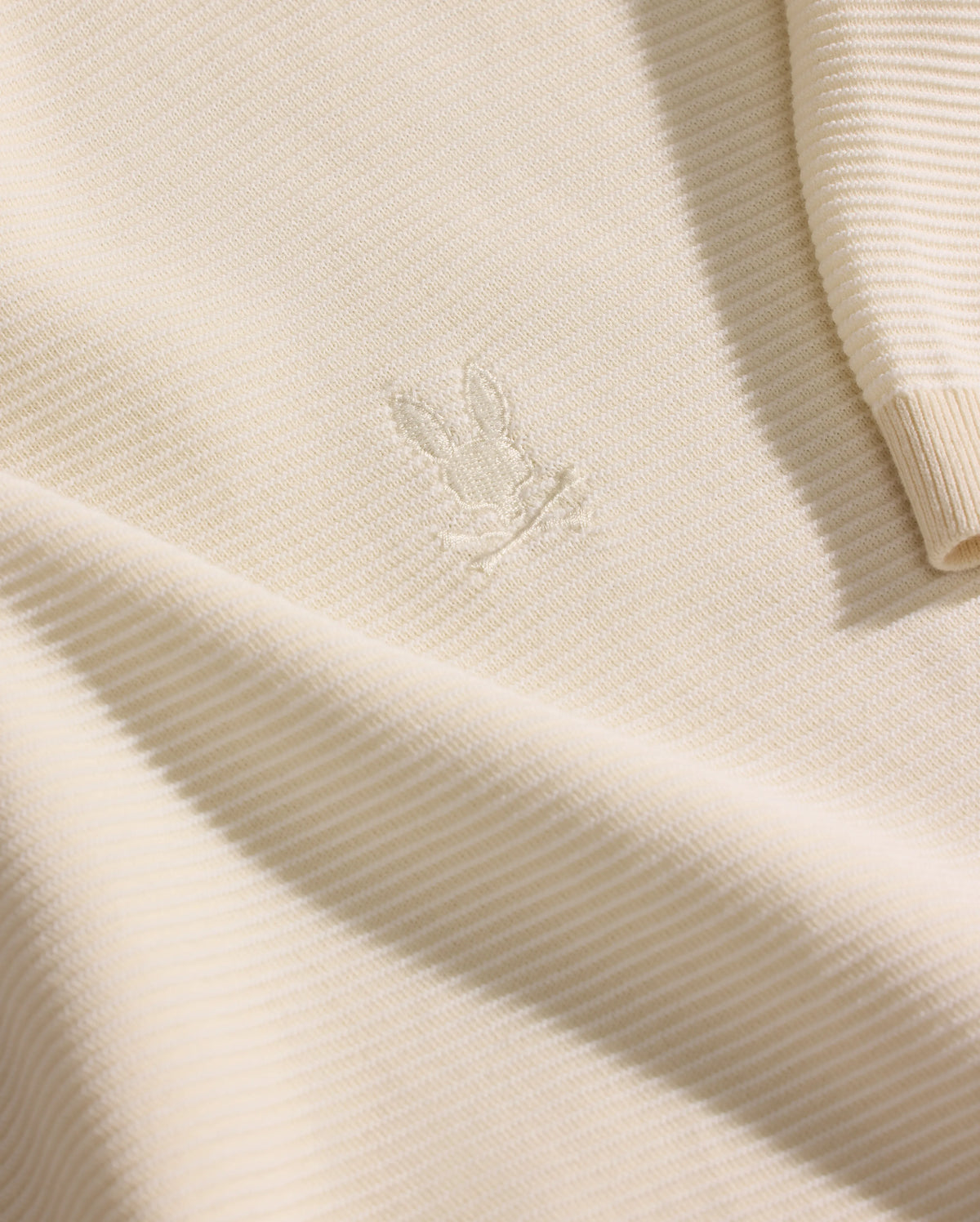 A close-up of the Psycho Bunny MENS MARIO STRIPED SWEATER T-SHIRT (MT0400009) in cream shows ribbed knit fabric with an embroidered bunny head on the chest and a partially visible sleeve highlighting the striped texture.