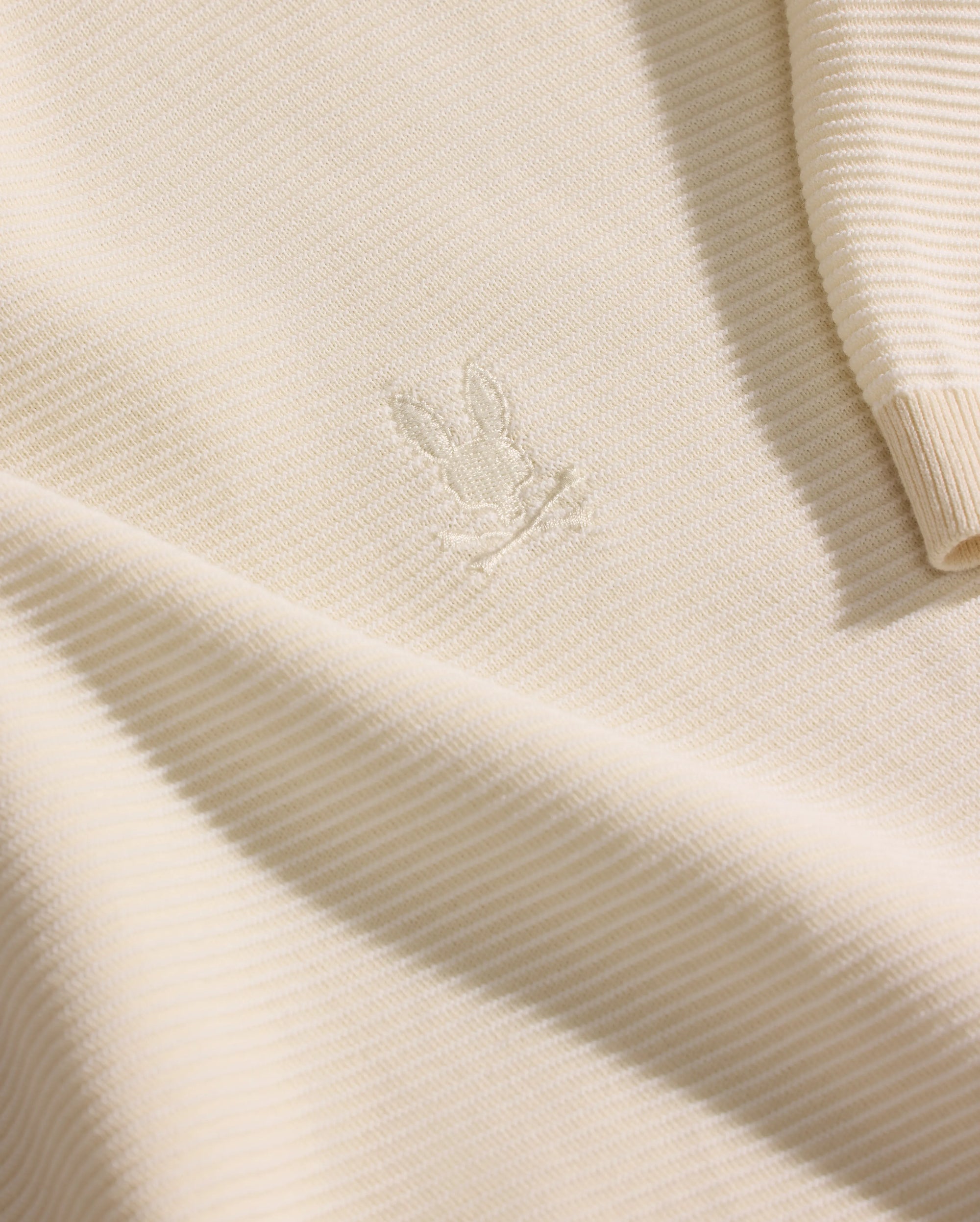 A close-up of the Psycho Bunny MENS MARIO STRIPED SWEATER T-SHIRT (MT0400009) in cream shows ribbed knit fabric with an embroidered bunny head on the chest and a partially visible sleeve highlighting the striped texture.