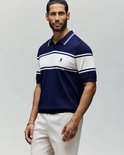 A man with short dark hair and a beard wears the Psycho Bunny MENS ZAVIAN STRIPE KNIT POLO (MT0400055) in navy and white stripes, paired with light shorts, standing against a plain light gray background.