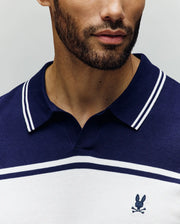 A man with a short beard wears the Psycho Bunny MENS ZAVIAN STRIPE KNIT POLO (MT0400055) in navy and white, featuring a small embroidered bunny and crossbones logo on the chest. Photo shows only his lower face and upper torso.