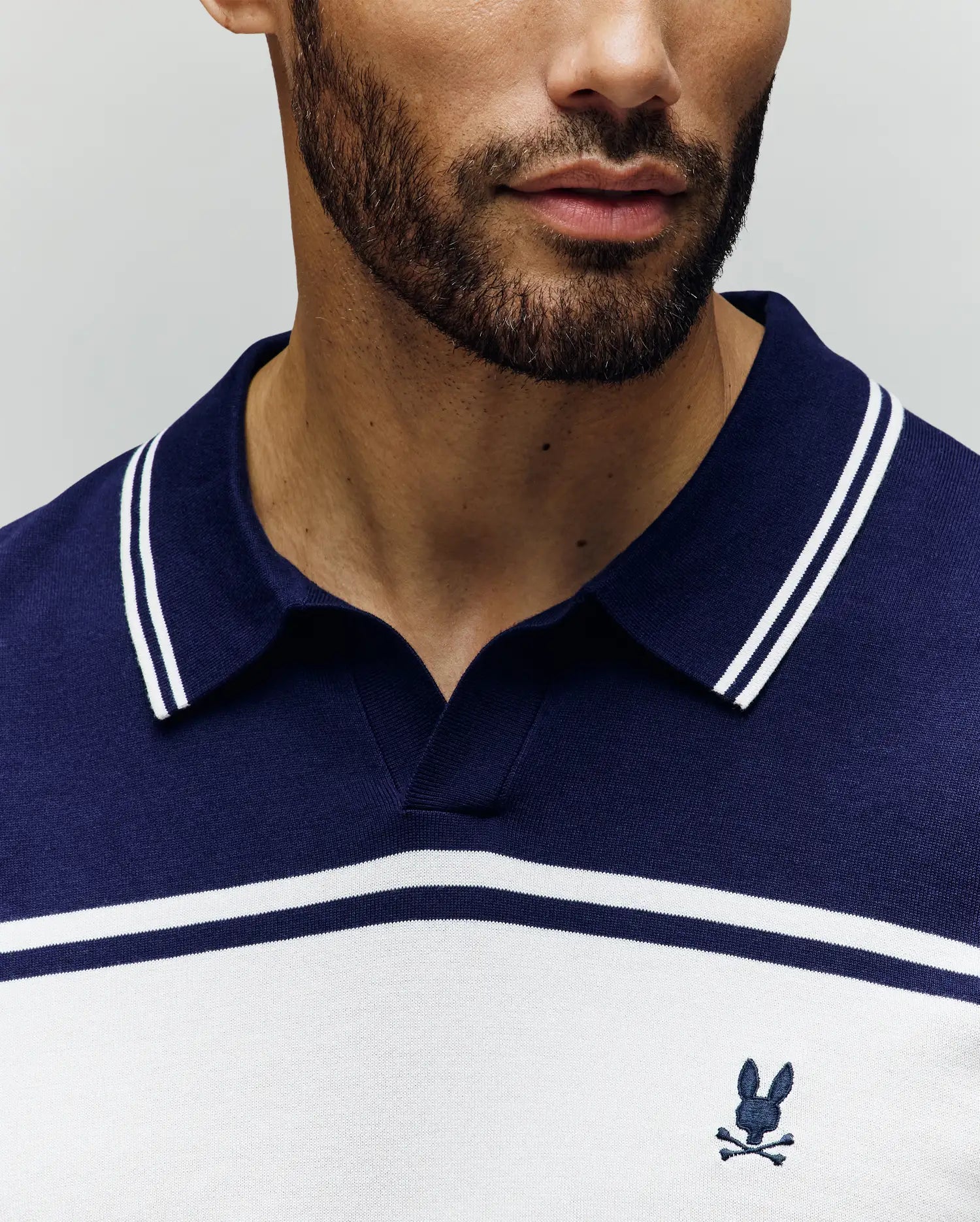 A man with a short beard wears the Psycho Bunny MENS ZAVIAN STRIPE KNIT POLO (MT0400055) in navy and white, featuring a small embroidered bunny and crossbones logo on the chest. Photo shows only his lower face and upper torso.
