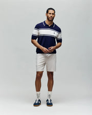 A man with short dark hair and a trimmed beard stands against a plain background, wearing the Psycho Bunny MENS ZAVIAN STRIPE KNIT POLO - MT0400055 in navy and white, light shorts, patterned socks, and navy sneakers. His hands are loosely clasped.