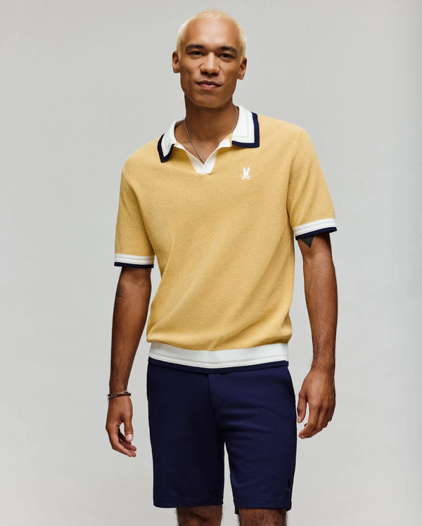 A man wearing the Psycho Bunny MENS CARTER JOHNNY COLLAR KNIT POLO - MT0400093 in yellow with white and navy trim and navy shorts stands against a light gray background, smiling slightly with his hands by his sides.