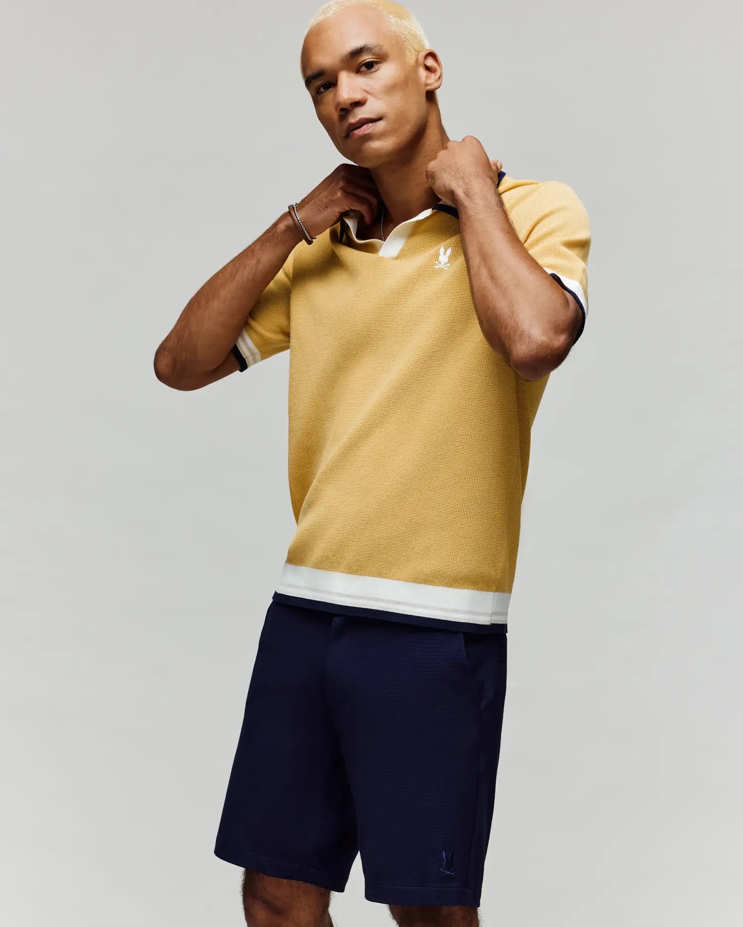 A person with short blond hair adjusts the collar of a Psycho Bunny MENS CARTER JOHNNY COLLAR KNIT POLO - MT0400093 in yellow, paired with navy shorts, standing against a plain light gray background.