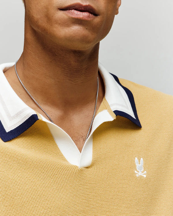 Close-up of a person wearing the Psycho Bunny MENS CARTER JOHNNY COLLAR KNIT POLO (MT0400093), a 100% cotton yellow shirt with white and navy trim, silver chain necklace, and an embroidered white bunny skull-and-crossbones logo. Face partially visible.
