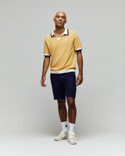 A person stands against a plain light background, wearing the Psycho Bunny MENS CARTER JOHNNY COLLAR KNIT POLO in yellow, paired with navy shorts, white logo crew socks, and white sneakers. Their look is relaxed and casual.