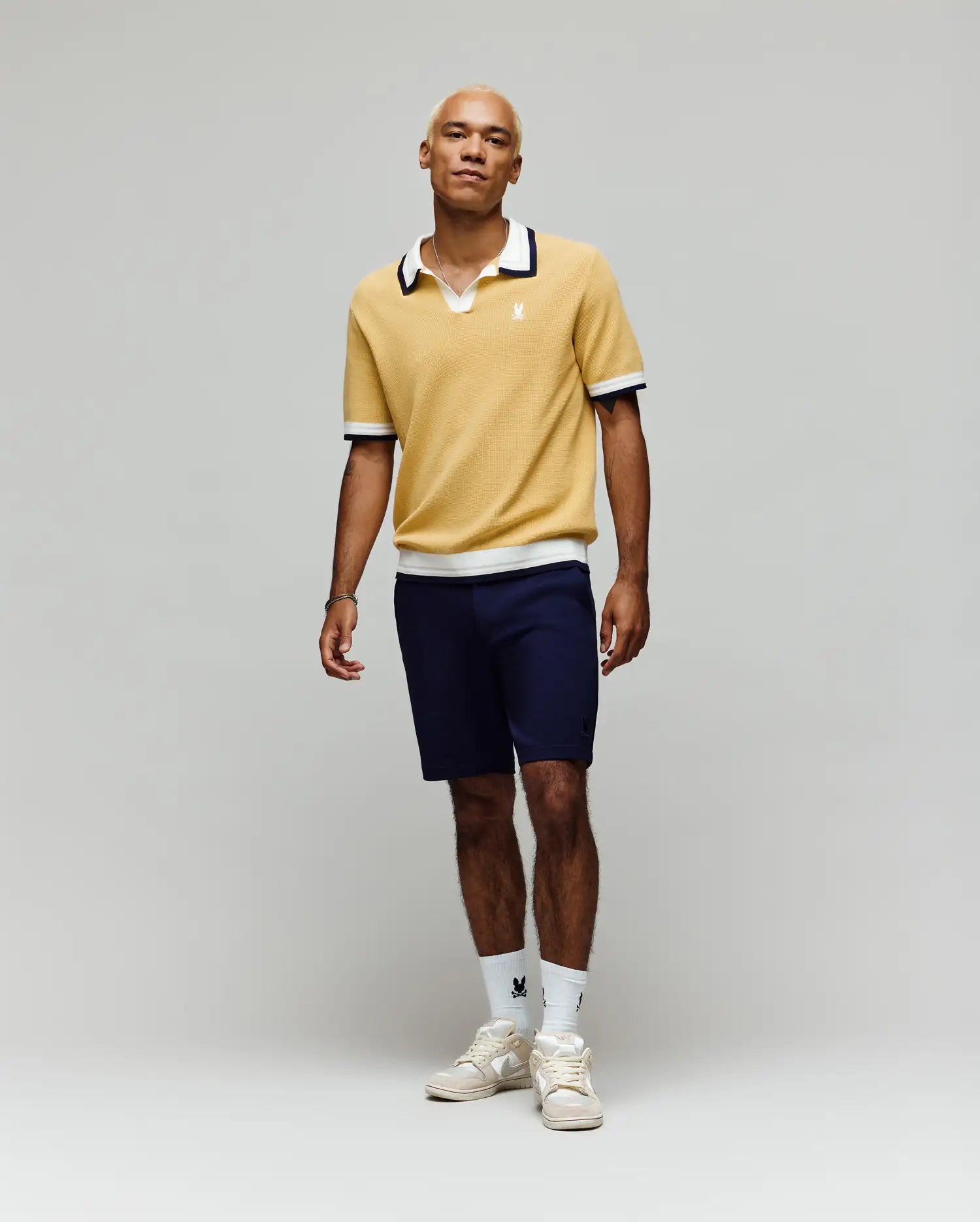 A person stands against a plain light background, wearing the Psycho Bunny MENS CARTER JOHNNY COLLAR KNIT POLO in yellow, paired with navy shorts, white logo crew socks, and white sneakers. Their look is relaxed and casual.