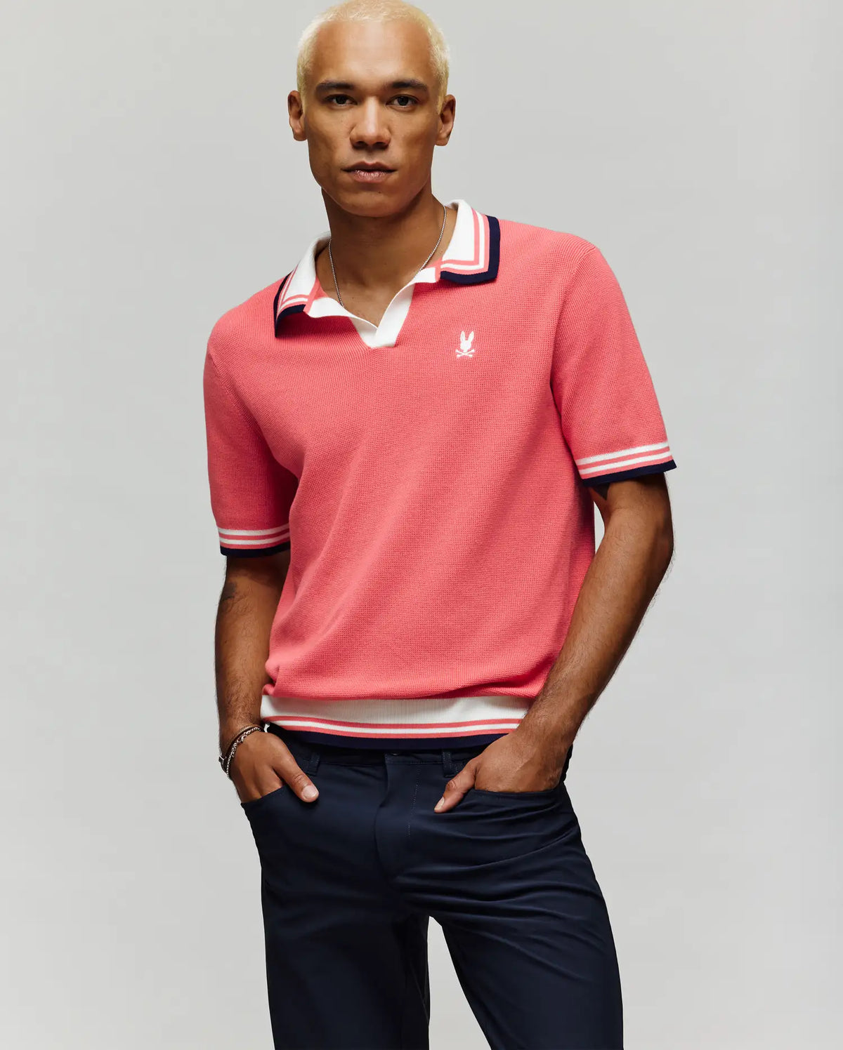 A man with short, bleached blond hair models the Psycho Bunny MENS CARTER JOHNNY COLLAR KNIT POLO - MT0400093, a 100% cotton pink polo with white and navy trim, paired with dark pants against a plain light background.
