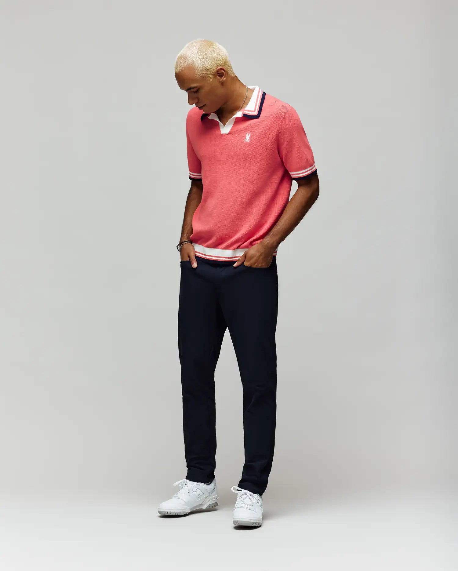 A person with short blond hair wears the Psycho Bunny MENS CARTER JOHNNY COLLAR KNIT POLO (MT0400093) in pink, paired with dark pants and white sneakers, standing against a plain light gray background and looking down with hands in pockets.